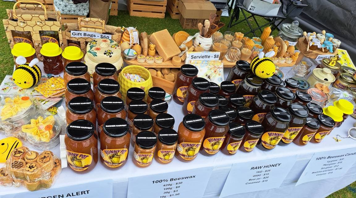 Market stall displaying Hunny Pot honey jars, beeswax candles, serviettes, gift bags, and handcrafted bee-themed items including crocheted bees and honey dippers